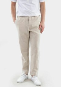 Beige Leinenhosen mit elastischem Bund, schmaler Passform und Seitentaschen, kombiniert mit weißen Sneakern vor einem einfarbigen Hintergrund.
