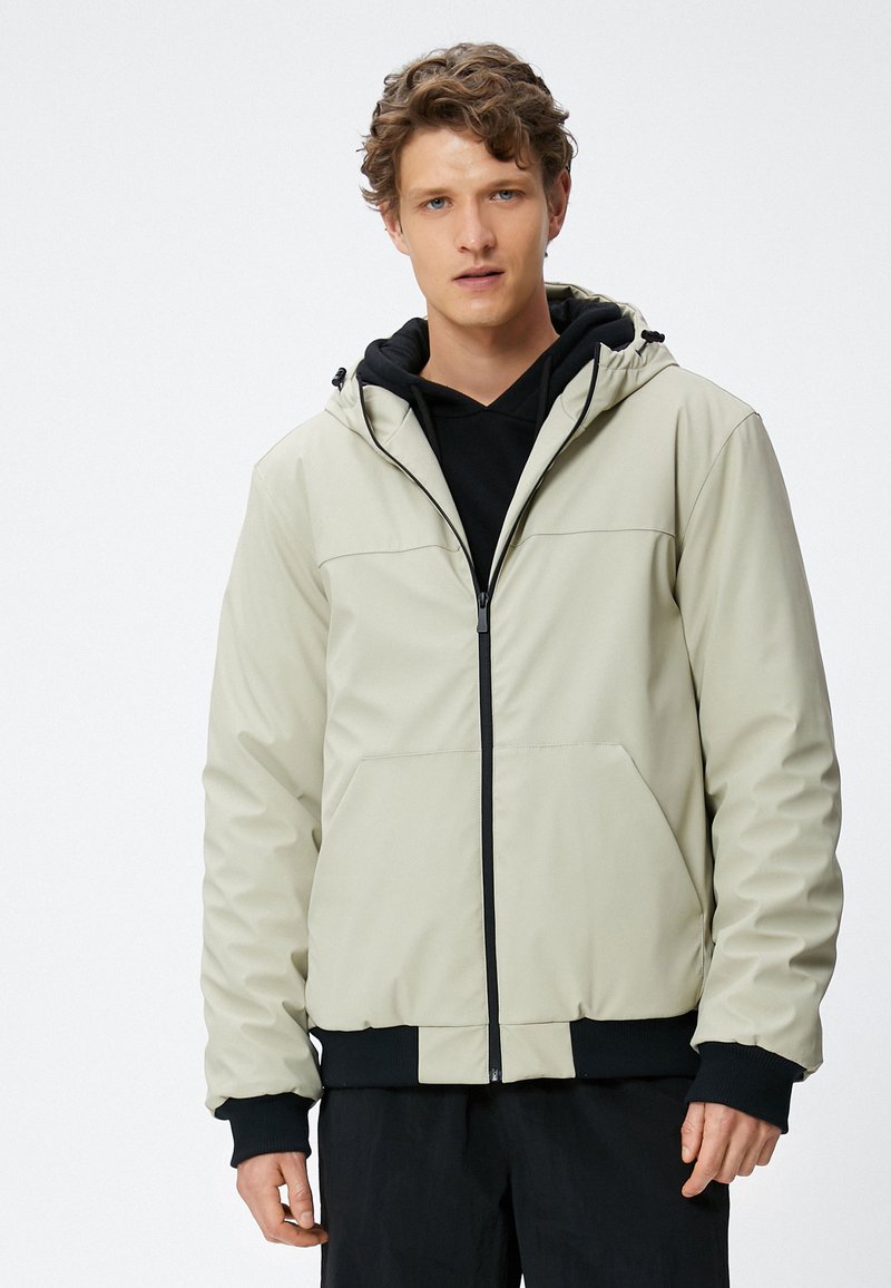 Licht beige capuchonjack met een ritssluiting aan de voorkant, zwarte accenten op de mouwen en onderkant, met een gladde textuur en twee voorzakken.