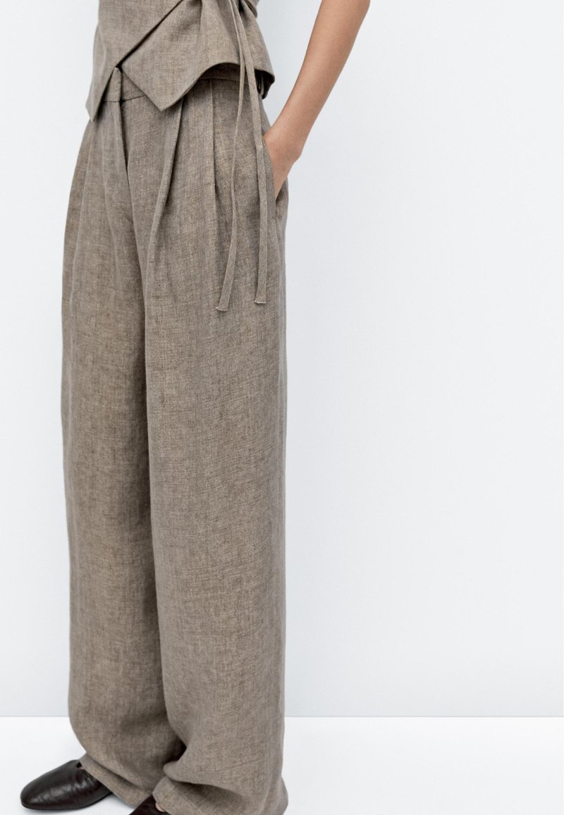 Lichtbruine linnen broek met een brede pijp, voorzien van een hoge tailleband en zijzakken. Touwtjes hangen van de tailleband voor aanpassing.