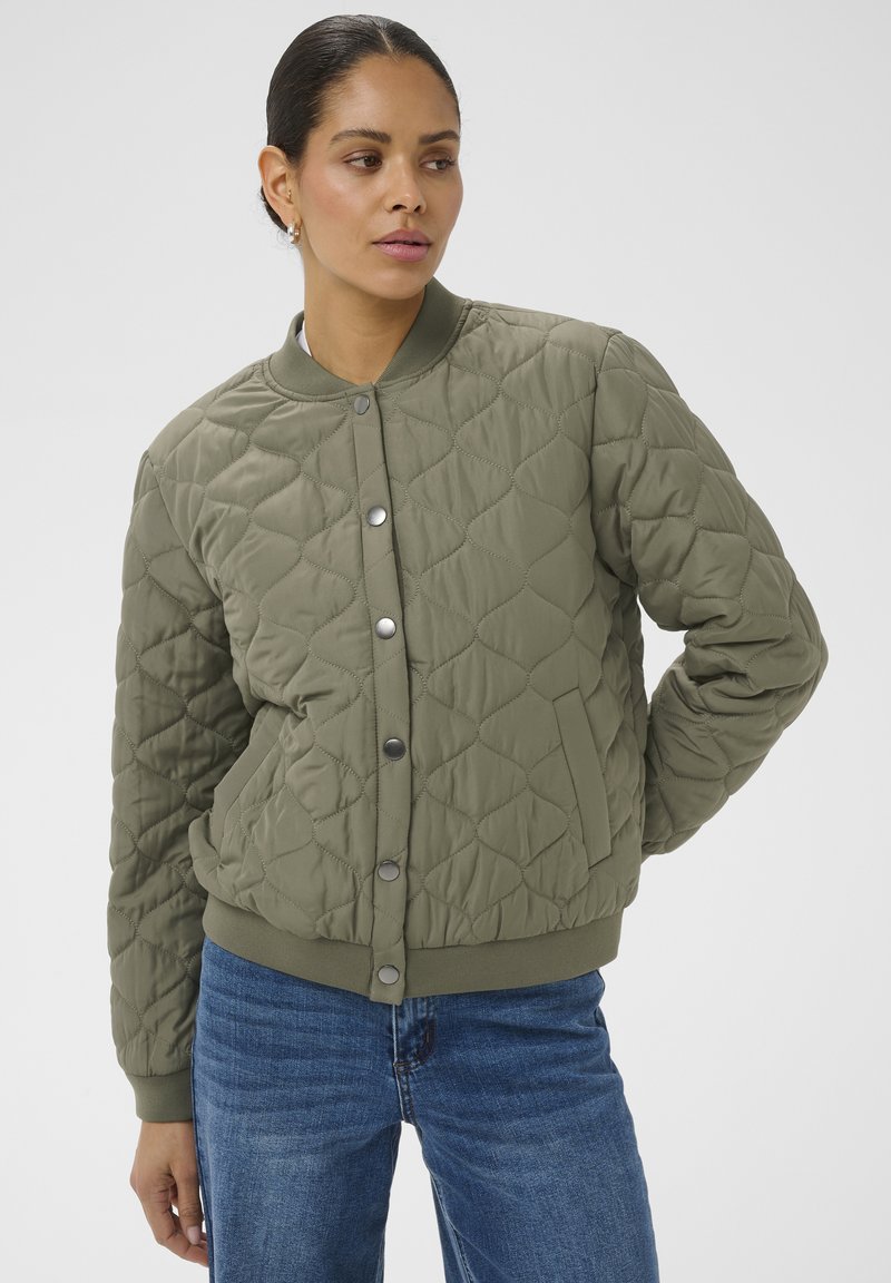 Femme portant une veste bomber matelassée vert olive avec des boutons-pression et un jean bleu, debout avec une main derrière le dos.