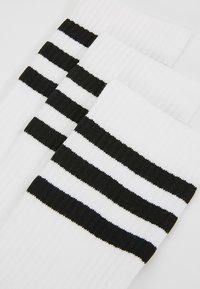 Weiße Socken mit drei horizontalen schwarzen Streifen an der Oberseite. Hergestellt aus einem gerippten, strukturierten Stoff für Flexibilität und Tragekomfort.