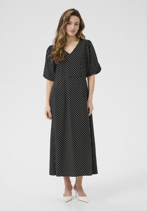 Zwarte maxi-jurk met polka dots, met een V-hals, korte pofmouwen en een vloeiende silhouet. Gekleed met witte puntenhakken.