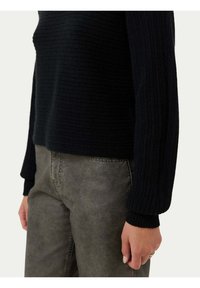 Svart ribbat stickat tröja med långa ärmar, i kombination med grå texturerade byxor. Den synliga handen vilar på midjan.