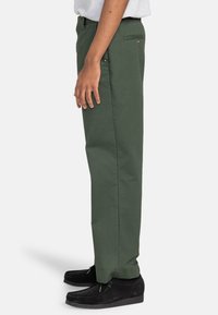 Pantalon en coton vert au design droit, avec poches latérales et une poche arrière. Associé à des chaussures noires, mettant en valeur une texture lisse.