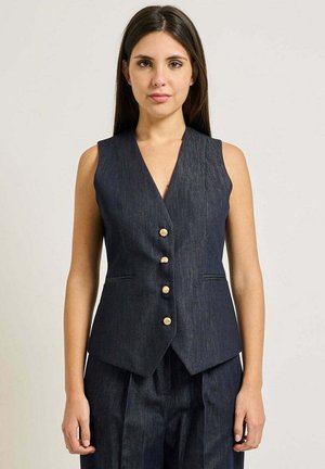 Donna con capelli lunghi e scuri che indossa un gilet di denim scuro senza maniche con bottoni dorati e pantaloni abbinati, in piedi su uno sfondo chiaro.