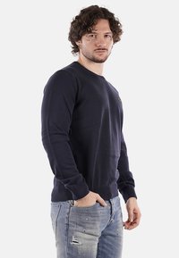 Felpa blu navy a girocollo in cotone con polsini e orlo a costine, indossata sopra jeans chiari strappati, caratterizzata da ricami sottili.