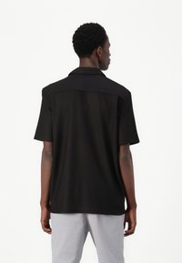 Cămașă neagră cu mânecă scurtă, cu un guler structurat și țesătură texturată. Partea din spate are un design solid și este asortată cu pantaloni de culoare deschisă.