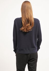 Pull bleu marine avec des rayures horizontales texturées, col rond, poignets et ourlet côtelés ; porté avec un pantalon noir, vu de dos.