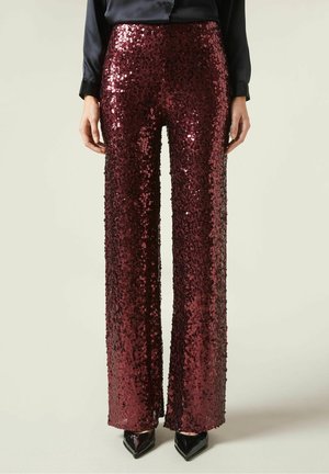 Kobieta w bordowych, szerokich spodniach z cekinami z wysokim stanem, w połączeniu z czarną koszulą na guziki i czarnymi spiczastymi butami.