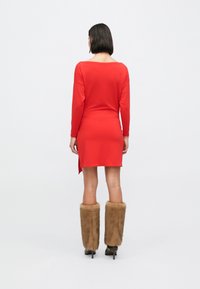 Czerwona sukienka z długim rękawem, z dekoltem w łódkę i asymetrycznym dołem. Noszona z brązowymi futrzanymi getrami i butami na obcasie. Prosty, dopasowany krój.
