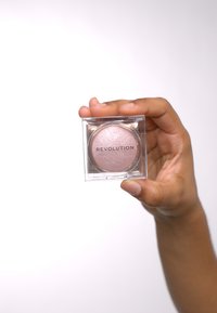 Revolution highlighter i en klar firkantet beholder. Myk rosa pulver med shimmer, sirkulær form, med et trykt merkenavn på forsiden.