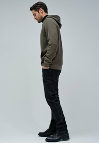 Felpa marrone con texture, dotata di tasca a marsupio, abbinata a jeans neri e scarpe nere, mostrata in profilo laterale su uno sfondo semplice.