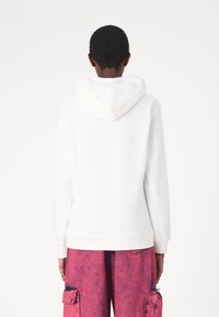 Sudadera blanca con un bolsillo canguro frontal y capucha ajustable, combinada con pantalones cargo rosas que cuentan con un bolsillo con cremallera y una textura gastada.