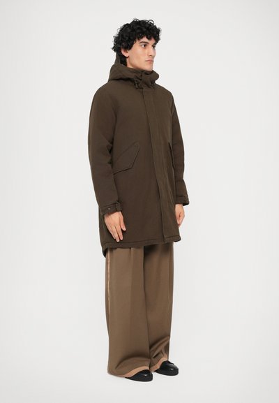Parka maronie cu glugă, având o croială dreaptă, închidere frontală, buzunare laterale și țesătură texturată. Asortată cu pantaloni largi maronii.