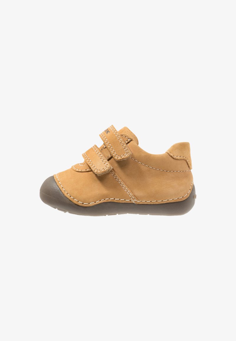 Chaussure d’enfant en daim beige avec deux brides Velcro, des surpiqûres blanches, et une pointe ainsi qu’une semelle en caoutchouc marron, vue de côté sur fond blanc.