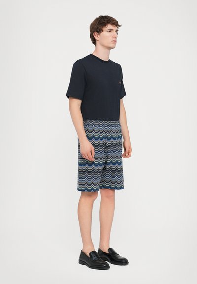 Marinblå bomullströja med korta ärmar, i kombination med mönstrade shorts med vågiga blå, gröna och vita ränder. Svarta loafers fulländar looken.