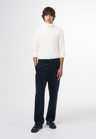 Weißes Rollkragenoberteil kombiniert mit dunkelblauen Cordhosen. Die Hosen haben einen Knopfverschluss und Vordertaschen. Schwarze Schuhe runden das Outfit ab.