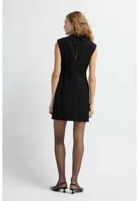 Schwarzes ärmelloses Kleid mit strukturiertem Design, das einen hohen Kragen mit Perlenverzierung und einen Reißverschlussverschluss hinten aufweist.
