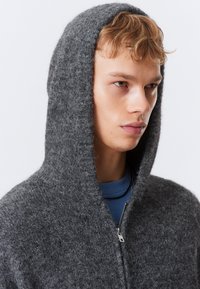 Joven con una sudadera gris de felpa con cremallera y capucha puesta, mirando hacia la derecha contra un fondo blanco liso.