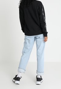 Zwarte sweatshirt met witte logoprint op de mouwen, gecombineerd met lichtblauwe opgerolde jeans en zwarte sneakers met witte zolen.