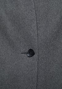 Šedá keprová látka s texturovaným povrchem; na levé straně má jeden černý knoflík, který přidává detaily do designu.