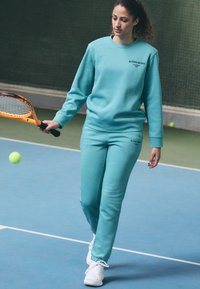 Una mujer con un sudadera turquesa y joggers a juego, sosteniendo una raqueta de tenis. Zapatos deportivos blancos, textura de tela suave, diseño minimalista.
