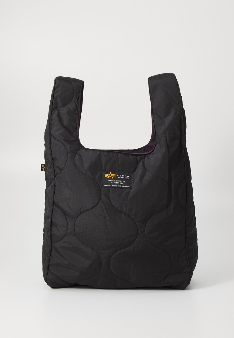 Črna prešita torba z veliko odprtino, ukrivljenimi oblikami na površini in oznako z rumenim in belim besedilom spredaj.