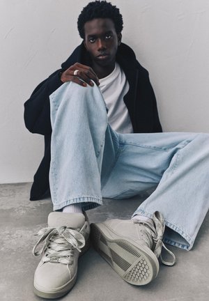 Tânăr cu păr creț și închis la culoare, purtând cămașă albă, jachetă neagră, blugi bleu deschis și adidași gri, stă pe podea de beton lângă un perete alb.