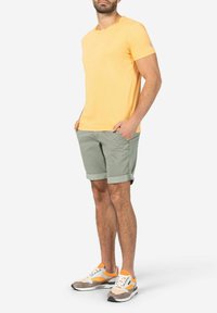 Gelbes T-Shirt mit kurzen Ärmeln, kombiniert mit grünen Shorts. Die Schuhmode hat graue, orangefarbene und weiße Akzente. Lässiges Sommeroutfit.