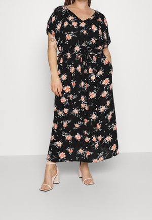 Femme portant une robe longue noire à fleurs avec manches courtes et ceinture à la taille, associée à des talons blocs ouverts rose clair.