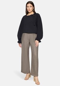 Zwarte blouse met gerimpelde mouwen en strikdetail, gecombineerd met lichtbruine wijde broekspijpen. Model draagt lichtbeige sandalen met blokhak.