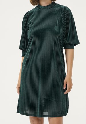 Femme portant une robe en velours vert foncé à rayures verticales, manches bouffantes mi-longues et un col haut à volants.