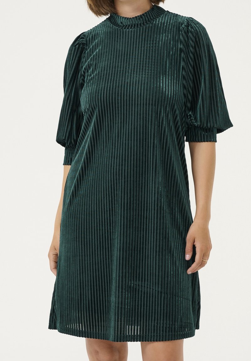 Femme portant une robe en velours vert foncé à rayures verticales, manches bouffantes mi-longues et un col haut à volants.
