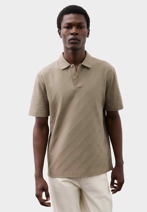 Man draagt een beige polo met korte mouwen met gestructureerde diagonale strepen en lichte broek tegen een effen achtergrond.