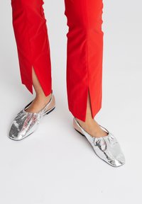 Zilverkleurige ballerina's met een gestructureerde afwerking, voorzien van een open achterkant en een ronde neus. Gecombineerd met rode broek met zij-splitten.