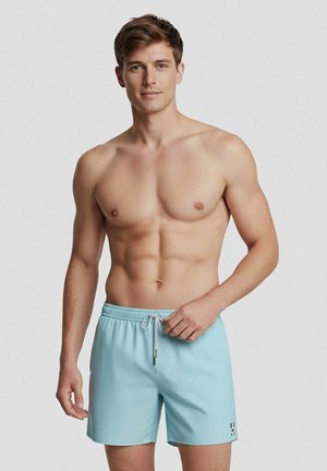 Jonge man zonder shirt die lichtblauwe zwembroek met wit trekkoord aanpast tegen een effen achtergrond.