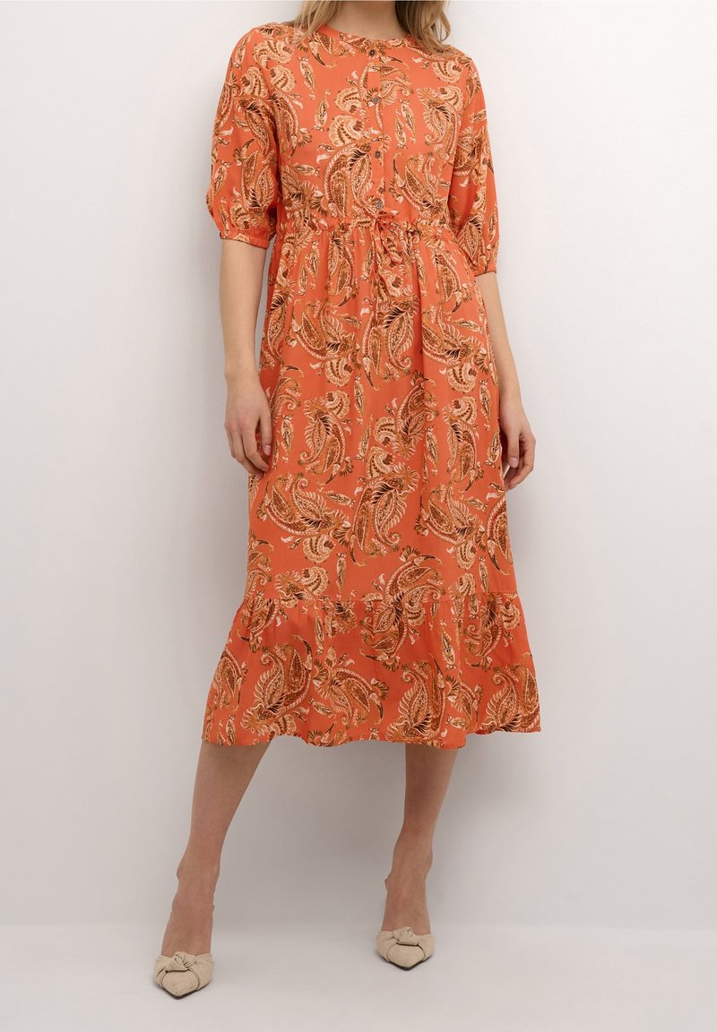 Femme portant une robe orange mi-longue avec des motifs paisley et des manches courtes, associée à des escarpins beiges à bout pointu.
