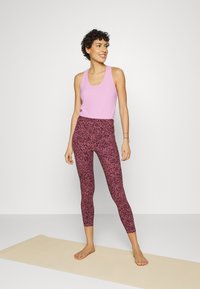 Camisola de alças rosa clara combinada com leggings justas com um padrão abstrato em rosa escuro e preto. Modelo está descalça em cima de um tapete de yoga.