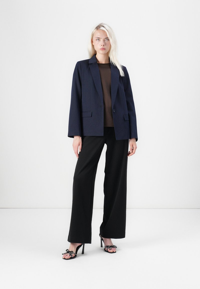 Blazer navy avec épaules structurées, associé à un haut marron, un pantalon noir à larges jambes et des sandales à talons hauts noires avec des nœuds.