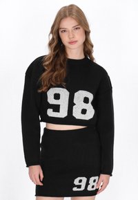 Fekete kötött, cropped pulóver fehér "98" felirattal az elején, amelyet egy hozzá illő fekete szoknyával párosítanak, amely alsó részén fehér "98" található.