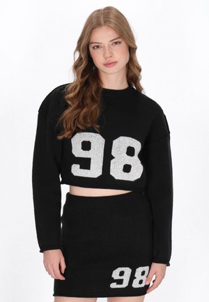 Schwarzer, gestrickter, kurzer Pullover mit weißem "98"-Druck auf der Vorderseite, kombiniert mit einem passendem schwarzen Rock, der unten einen weißen "98"-Druck aufweist.