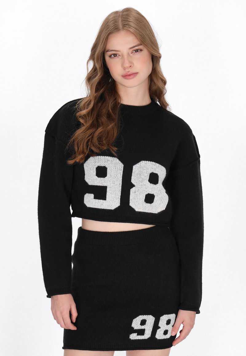 Fekete kötött, cropped pulóver fehér "98" felirattal az elején, amelyet egy hozzá illő fekete szoknyával párosítanak, amely alsó részén fehér "98" található.