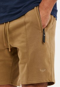 Shorts en polaire marron avec taille à cordon, poche zippée latérale et motif brodé sur la jambe. Texture douce, coupe décontractée.