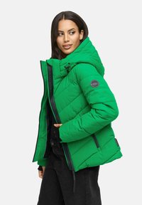 Femme portant une veste rembourrée verte vif avec capuche, un pantalon noir, regardant sur le côté contre un fond uni.