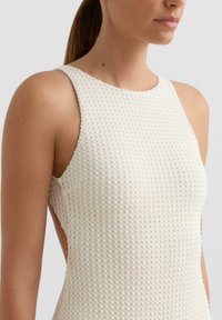 Bezruké krémové šaty s vaflovou textúrou, vysokým golierom a otvorenými stranami; priliehavý strih zvýrazňuje siluetu.