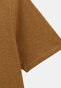 Manche courte d'un T-shirt en coton texturé marron avec des coutures visibles sur l'ourlet, sur un fond clair.