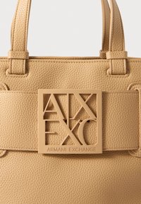 Beżowa torba skórzana o fakturze z wyszywanymi paskami i dużą kwadratową plakietką z logo Armani Exchange z przodu.
