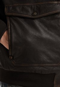 Brun læderjakke med tekstureret overflade, en frontlomme lukket med en metaltrykknap og ribbet stof ved kanten.