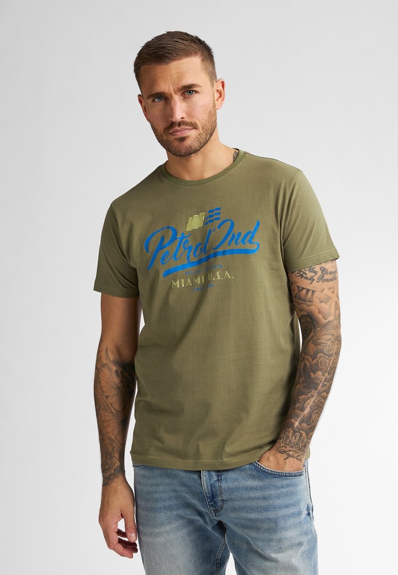 T-shirt en coton vert olive avec un texte imprimé bleu et or "Petrol Ind Miami U.S.A." Le modèle porte le t-shirt avec un jean bleu clair.