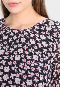 Haut fleuri avec fond noir, présentant des motifs de fleurs blanches et rouges, un col plissé et un tissu léger et transparent.